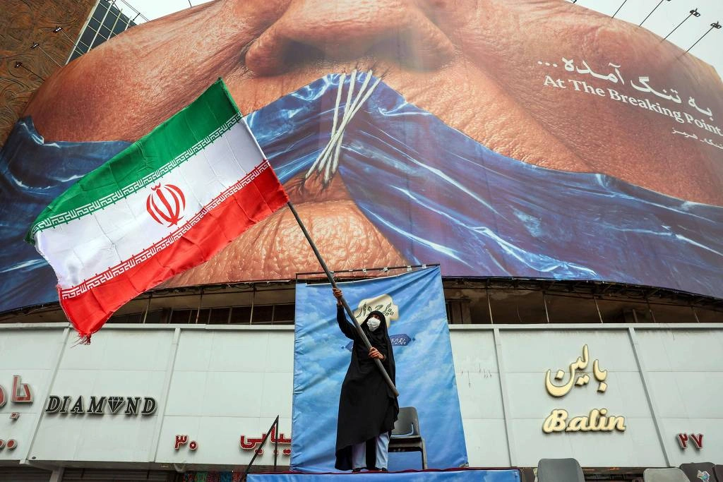 Mulher segura bandeira do Irã em frente à outdoor em Teerã que representa o presidente dos EUA, Donald Trump, e o fechamento do estreito de Hormuz