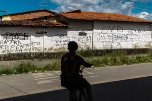 Pessoa com mochila sentada em bicicleta na sombra observa muro branco com várias pichações em preto. O muro tem telhado de cerâmica e está em área urbana com céu parcialmente nublado ao fundo.  Pichações mostram ordens e siglas de facção criminosa em rua de Maranguape, na região metropolitana de Fortaleza (CE); município teve a maior taxa de homicídios estimados no país, com 87,2 casos para cada 1 (Foto: Jarbas Oliveira)