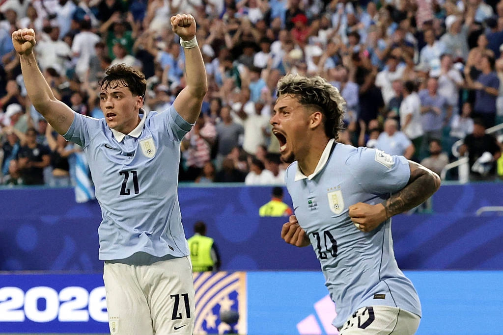 Dois jogadores da seleção uruguaia vestindo camisa azul clara e calção branco comemoram gol. Um levanta os braços e o outro grita com punhos cerrados. Torcida ao fundo aplaude em arquibancada cheia. Vinãs e Maxi Araújo celebram o gol de empate do Uruguai na partida em Miami pela Copa do Mundo
